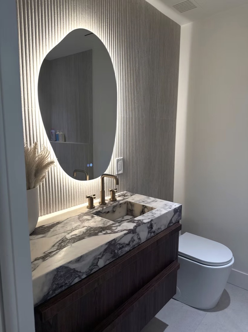 Powder room remodel in St. Petersburg, FL with a stone waterfall vanity, backlit oval mirror and fluted wall panel